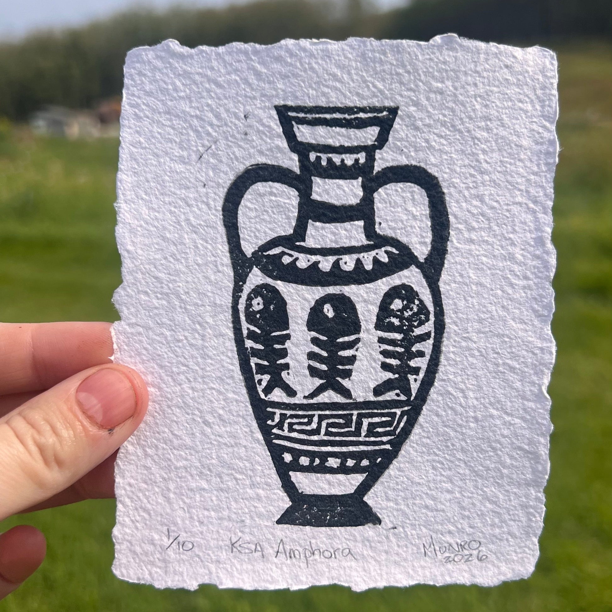 A hand holds a square piece of textured white paper with a black ink illustration of an ornate vase, photographed outdoors against a blurred green field and blue sky.
